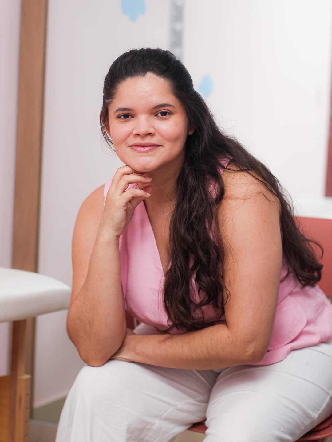 Retrato de Gleiciane Abreu sentada em seu espaço de atendimento, com blusa rosa e a mão apoiada no rosto